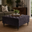 A dark grey tufted velvet ottoman with turned legs sits in a styled living room setting on a woven rug, with a sofa, sideboard, and decorative plants in the background.