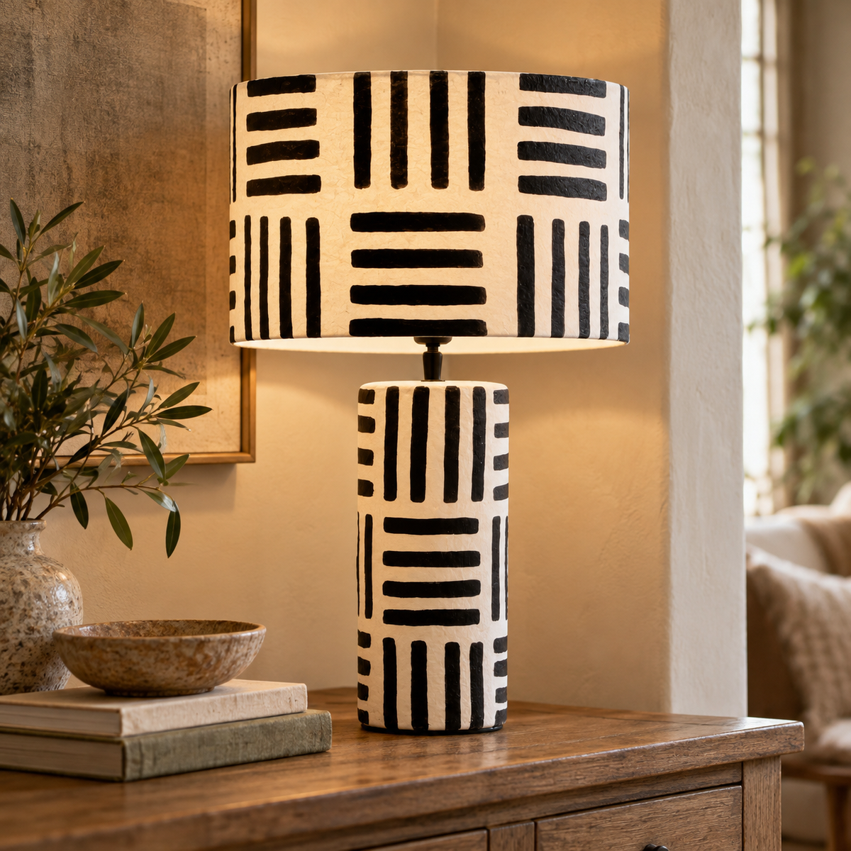 Luma Black And White Papier Mache Table Lamp with Geometric Lines lit on a wooden console beside olive branches