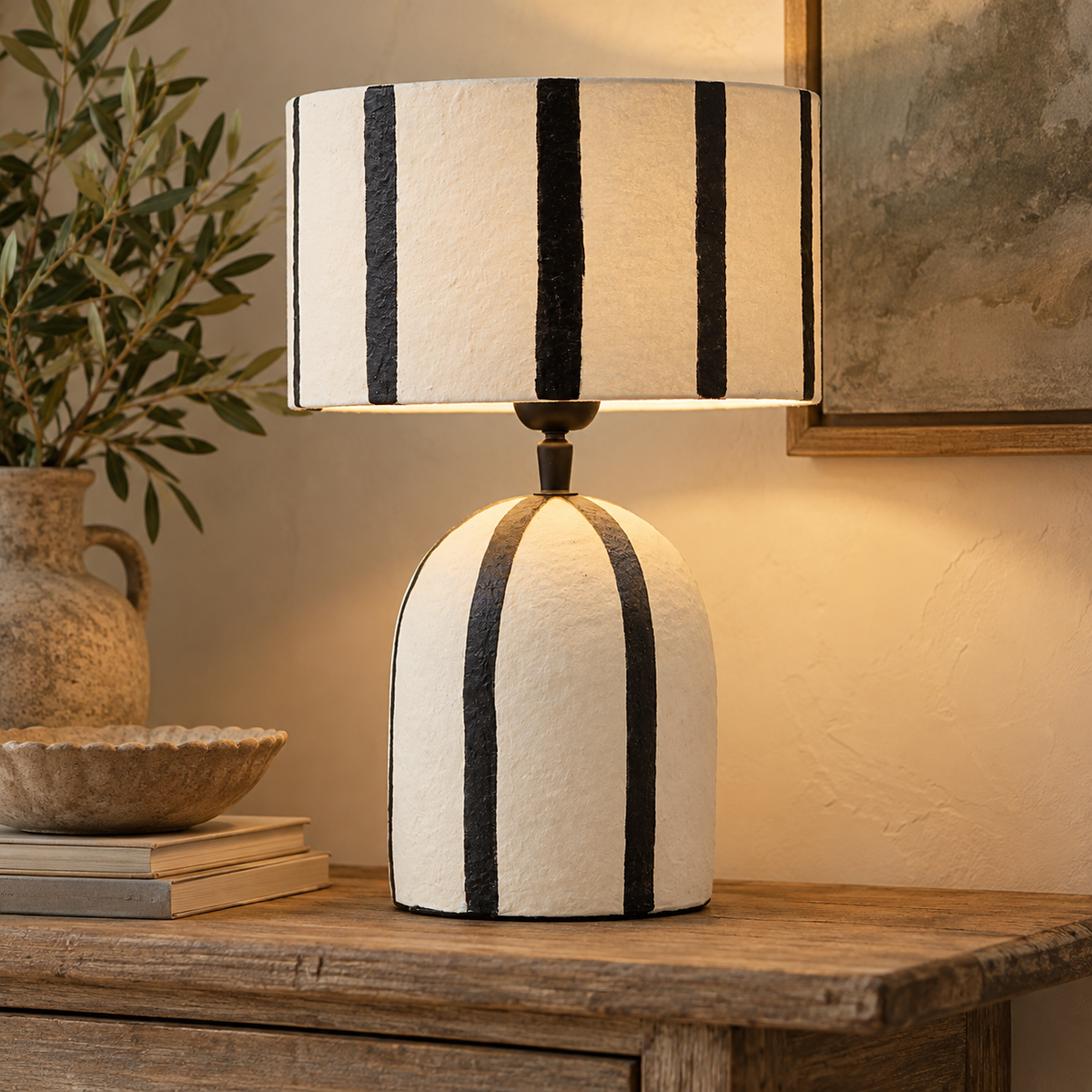 Luma Black And White Stripe Papier Mache Table Lamp lit on a wooden console with olive branches and books
