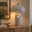 Luma Black And White Stripe Papier Mache Table Lamp With Dome Shade lit on a wooden console beside olive branches