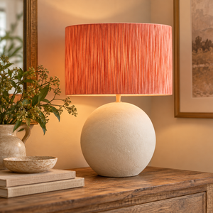 Luma Natural Papier Mache Table Lamp With Rust Raffia Shade lit on a wooden console beside greenery and books