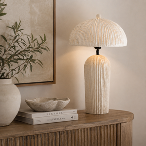 A textured cream table lamp with a ribbed cylindrical base and a matching ribbed dome shade, shown lit on a wooden console table in a styled interior setting.