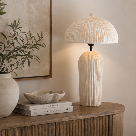 A textured cream table lamp with a ribbed cylindrical base and a matching ribbed dome shade, shown lit on a wooden console table in a styled interior setting.