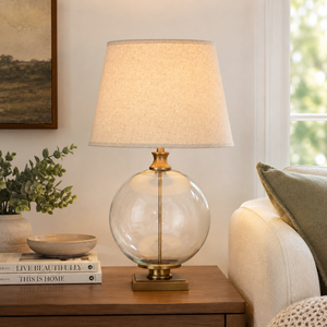 Table lamp with a glass base and beige shade on a wooden table in a living room setting.