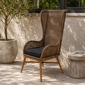 A tall outdoor wicker armchair with a dark cushioned seat and light wood legs, placed on a patio beside a potted plant and stone side table.
