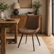 A brown upholstered dining chair with vertical channel tufting and black metal legs, shown in a styled dining room setting next to a rustic wooden table.