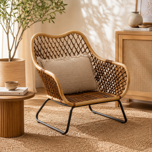 Marano black iron frame woven chair with a pillow in a cosy room setting with a plant and side table.
