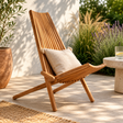 Murano wooden lounge chair with a pillow on a patio with plants and a table in the background