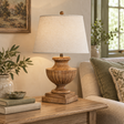 Marbelle Table Lamp With Linen Shade lit on a wooden side table beside a sofa with cushions and olive branches