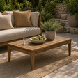 A rectangular wooden coffee table in a warm medium brown tone, styled on a beige rug in an outdoor patio setting. The table is decorated with a white bowl of green apples, a glass of water, and a potted green plant.