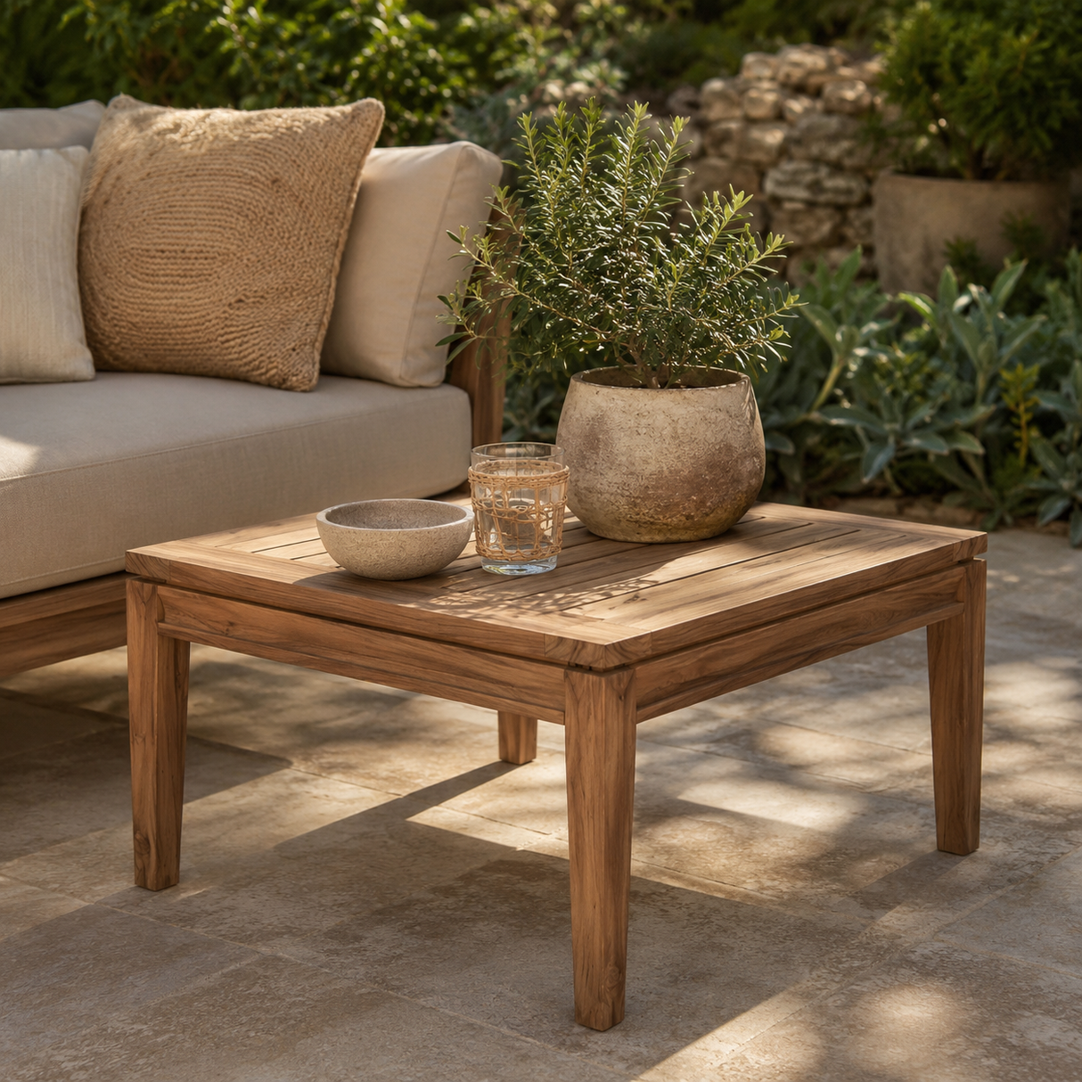 A square wooden outdoor coffee table in a warm medium brown tone, styled with a small bowl, a glass, and a potted green plant. It is positioned next to a beige outdoor sofa with textured pillows in a garden patio setting.