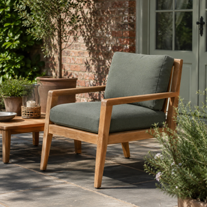 A wooden outdoor armchair with thick sage green cushions sits on a stone patio in a garden setting next to a matching wooden side table.