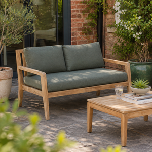 Outdoor wooden loveseat with thick sage green cushions on a stone patio, next to a matching wooden coffee table in a garden setting.