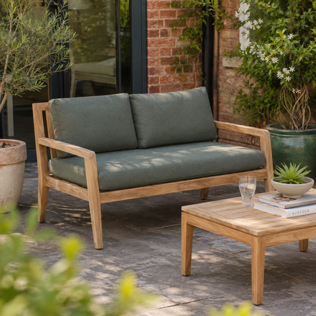 Outdoor wooden loveseat with thick sage green cushions on a stone patio, next to a matching wooden coffee table in a garden setting.