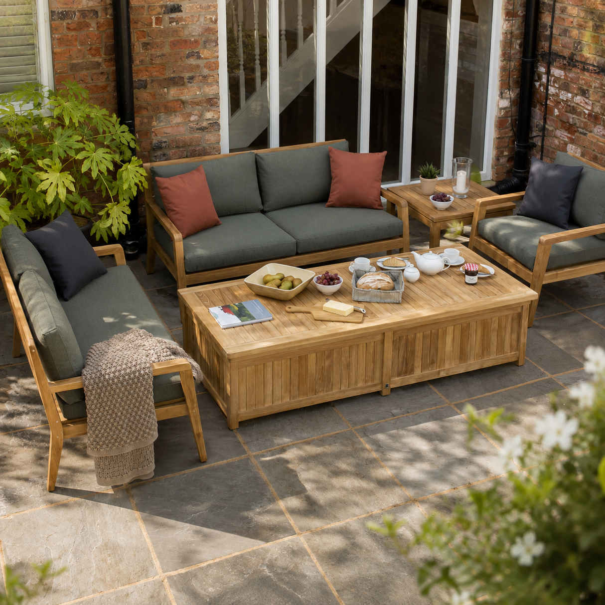 A 4-piece outdoor patio furniture set arranged on a stone paved terrace. The set includes a wooden coffee table, a wooden 3-seater sofa with dark green cushions and rust-colored pillows, a wooden armchair with dark green cushions and a dark pillow, and a small wooden side table.