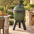 Moddern Kamado Barbecue in olive green on its black aluminium legs, on a sunlit stone terrace beside a laid garden table, olive trees and a log store in a dry stone wall