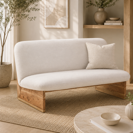 Two seater cream linen armless sofa with wooden base in a room with a woven rug, plant and decor items