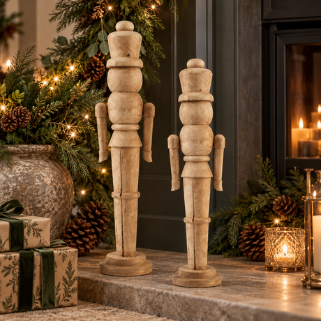 Small and large natural wooden nutcrackers on a hearth beside a lit fire, with presents, pinecones and candles