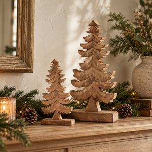 Small and large natural carved wooden trees on a mantel with pine sprigs, pinecones and a candle