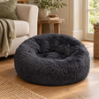 Navy Faux Fur Donut Dog Bed with long shaggy fur on a wooden floor beside a cream armchair and a green throw