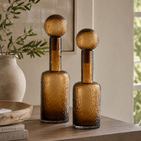 Two amber glass decorative bottles with textured hammered finishes and rounded stoppers, displayed on a wooden table in a styled interior setting.