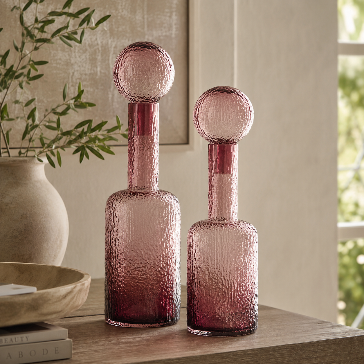 Two pink textured glass decanters with spherical stoppers, displayed on a wooden table in a styled interior setting.