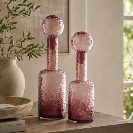 Two pink textured glass decanters with spherical stoppers, displayed on a wooden table in a styled interior setting.