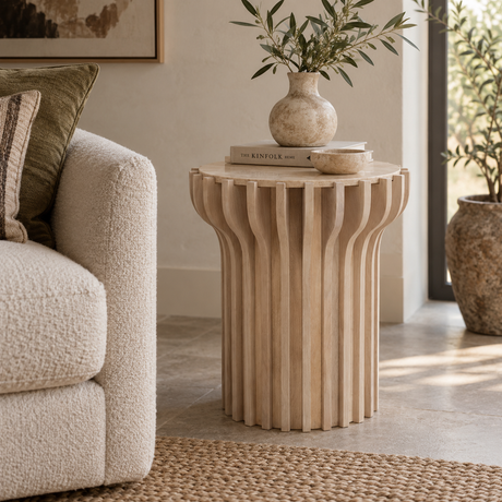 A light natural wood slatted side table in a styled living room setting, styled with a ceramic vase and books on top.