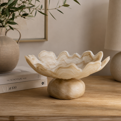A decorative bowl with a wavy, scalloped rim and a rounded pedestal base, shown on a wooden table in a styled interior setting with books, a vase, and a lamp.
