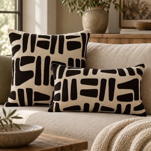Square and rectangular Elora Natural And Black Embroidered Cushions on a linen sofa with a knitted throw, beside a stone vase of olive branches