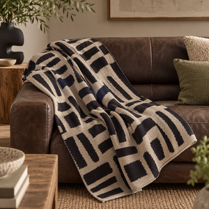 Elora Natural And Black Throw draped over a brown leather sofa with olive and boucle cushions, beside a rustic wooden table and jute rug