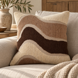 Elora Natural Assorted Colour Cushion with brown and sand wave bands on a cream sofa with a knitted throw, beside olive branches in a stone vase