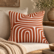 Elora Rust And White Cushion with rust arches on a linen sofa with a knitted throw, beside a wooden side table and olive branches
