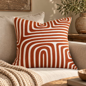 Elora Rust And White Cushion with rust arches on a linen sofa with a knitted throw, beside a wooden side table and olive branches