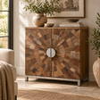 A rustic wooden sideboard cabinet with a circular sunburst pattern made from various wood tones on the doors, featuring a round silver metal handle and a silver metal frame. The cabinet is styled in a cosy living room setting with books, bowls, and potted plants.