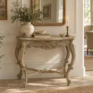 An ornate console table in a distressed beige finish, styled with a ceramic vase of greenery, books, and a candle in a bright, elegant room.