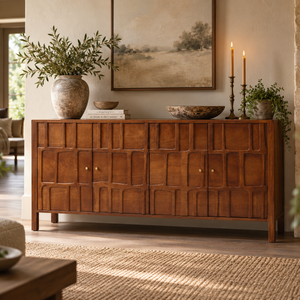 A warm medium-brown wooden sideboard with carved geometric panel details and small brass knobs, styled in a living room with a large vase of greenery, candles, bowls, and framed wall art.