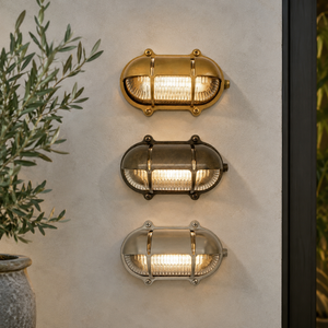 Three outdoor wall sconces in brass, bronze, and silver finishes mounted vertically on a textured beige wall, with a potted olive plant on the left.