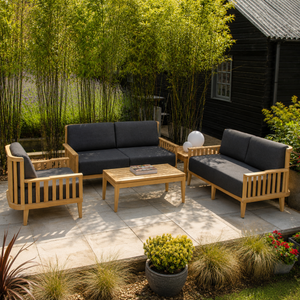A 4-piece outdoor patio furniture set with light natural wood frames and dark charcoal gray cushions, arranged on a stone patio in a garden setting with bamboo and a black wooden cabin in the background.