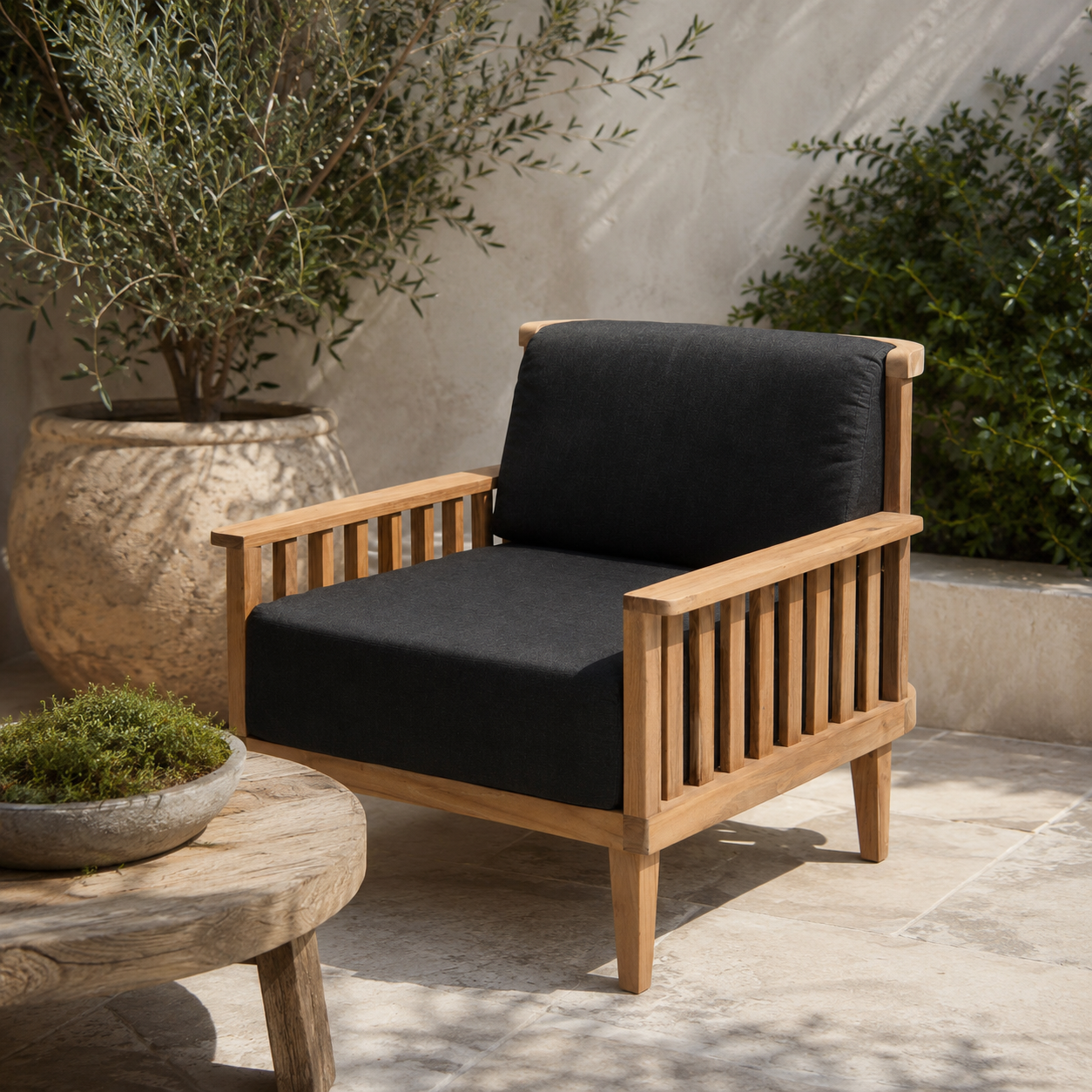 A wooden outdoor armchair with black cushioned seat and back cushions, placed on a stone patio with potted plants and a small round table nearby.