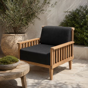 A wooden outdoor armchair with black cushioned seat and back cushions, placed on a stone patio with potted plants and a small round table nearby.