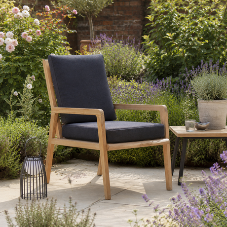 A wooden outdoor armchair with dark grey cushions sits on a stone patio in a lush garden setting, next to a small side table.