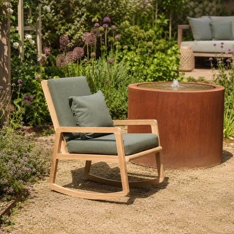 A wooden outdoor rocking chair with sage green cushions and a matching pillow, positioned next to a round rusted metal fire pit table in a landscaped garden patio setting.