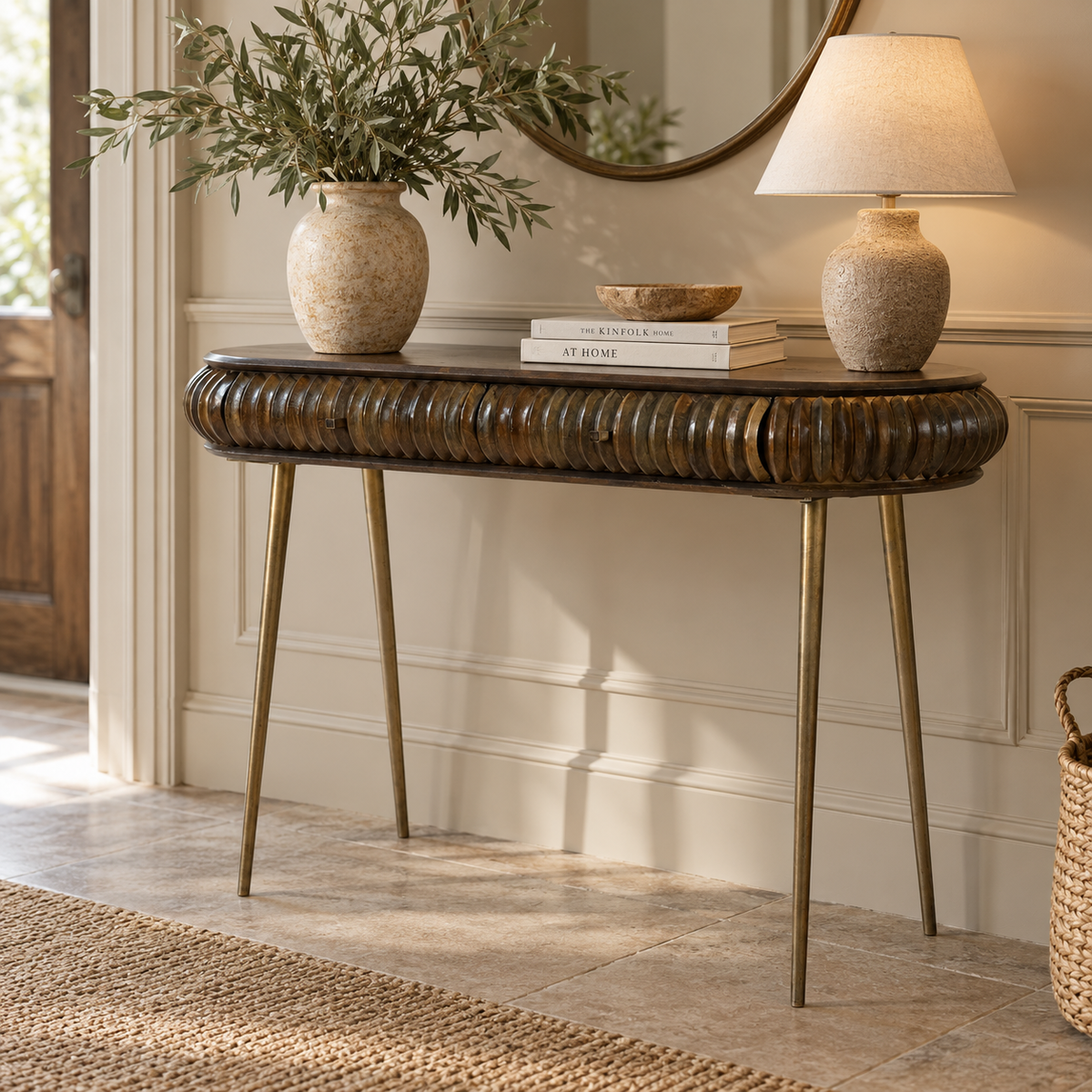 A console table with a dark brown ribbed wood top and brass legs, styled in a bright entryway with a vase of greenery, books, a lamp, and a mirror.