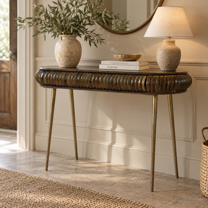 A console table with a dark brown ribbed wood top and brass legs, styled in a bright entryway with a vase of greenery, books, a lamp, and a mirror.