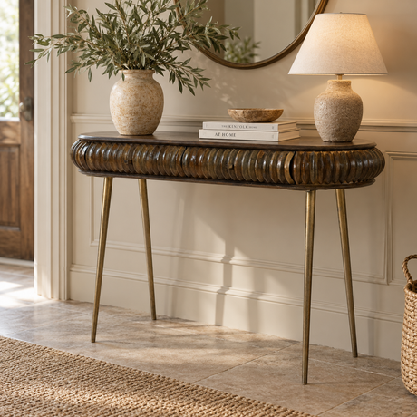 A console table with a dark brown ribbed wood top and brass legs, styled in a bright entryway with a vase of greenery, books, a lamp, and a mirror.