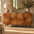 A mid-century modern wooden sideboard cabinet with four sunburst-patterned doors and tapered legs, styled in a warm, sunlit room with a lamp, mirror, and vases.