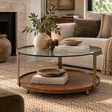 A round two-tier coffee table in a styled living room. The table features a warm medium-brown wood lower shelf, a clear glass upper surface, and a brass metal frame.
