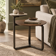 A round nesting side table set in a cozy living room. The set includes a smaller round table with a light stone top and a larger round table with a dark wood top, both supported by dark metal U-shaped legs.
