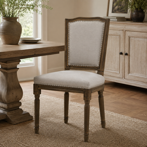 An upholstered wooden dining chair with a light beige fabric seat and backrest, featuring silver nailhead trim, shown in a dining room setting next to a rustic wooden table.